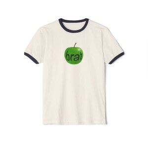 Può includere: Una t-shirt ringer color crema con bordi blu navy scuro sul colletto e sui polsini delle maniche. Al centro del petto è presente una grafica di una mela verde brillante con la parola "brat" scritta in minuscolo nero sopra il frutto.