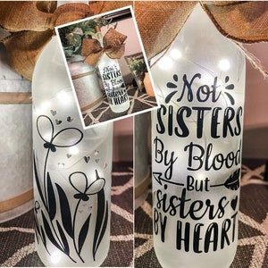 May include: A white frosted glass bottle with a string of lights inside. The bottle has a black vinyl decal with the text "Not Sisters By Blood But Sisters By Heart" and a small feather design. The bottle is decorated with a black floral design.