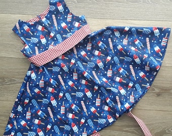 May include: A two-piece outfit featuring a blue fabric with a popsicle and star pattern. The top has a red and white gingham trim. The skirt is flared with a matching pattern and gingham tie.