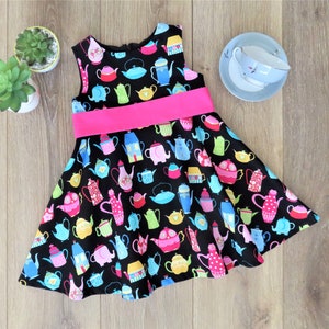 May include: A sleeveless black dress with a vibrant teapot pattern in various colors. The dress features a bright pink waistband and a flared skirt. A small teacup and saucer set is visible in the background.
