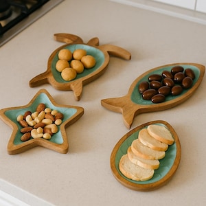 Puede incluir: Un conjunto de bandejas de madera para servir en varias formas: una tortuga, un pez, una estrella y un óvalo. Cada bandeja tiene un interior turquesa y está llena de diferentes alimentos, incluyendo nueces, rebanadas de pan y caramelos. Las bandejas están sobre una encimera clara.