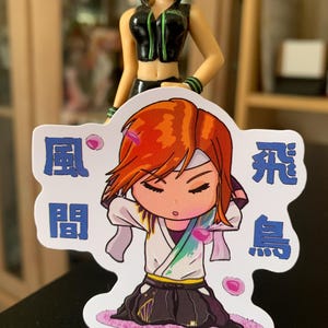 Può includere: Un adesivo con un personaggio dei cartoni animati con i capelli rossi, che indossa un abito bianco e nero, e il testo giapponese "風間 飛鳥". L'adesivo ha un bordo bianco ed è decorato con petali di fiori rosa.