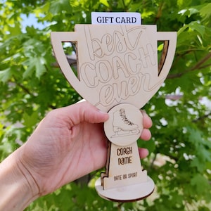 May include: Wooden trophy shaped like a cup with the text "best coach ever" engraved on it. The trophy has a small round cutout with a figure skate inside. The base of the trophy has the text "coach name" and "date or sport".