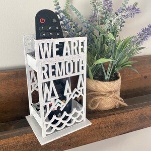 May include: White wooden remote control holder with the text "We are always looking for the remote" and a mountain and wave design. A black remote control is inside the holder.
