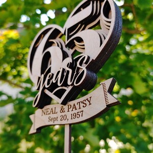 Pu&ograve; includere: Un topper per torta di anniversario in legno a forma di cuore con i numeri 65 e la parola "anni" in un carattere decorativo. Uno striscione sottostante recita "Neal & Patsy Sept 20, 1957".