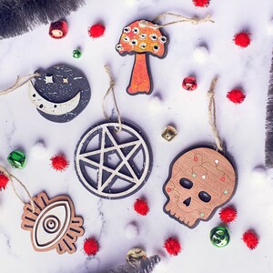 May include: A set of six wooden ornaments with various spooky and whimsical designs. The ornaments feature a crescent moon, a pentagram, a skull with lights, an eye, a mushroom, and a star. The ornaments are painted in black, white, and red.