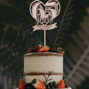 Pu&ograve; includere: Un topper per torta di legno a forma di cuore con il numero 65 e le parole "years" e "Neal & Patsy Sept 20, 1957" su uno stendardo. Il topper &egrave; su una torta a due piani decorata con fragole e mirtilli.