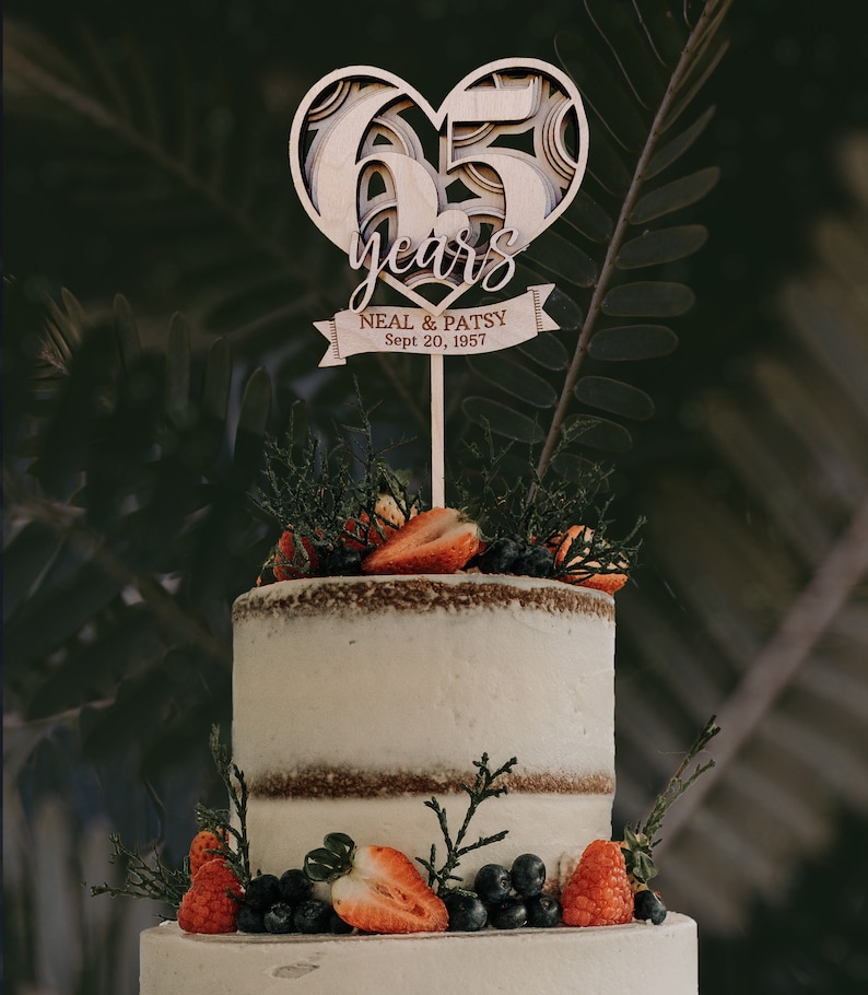 Pu&ograve; includere: Un topper per torta di legno a forma di cuore con il numero 65 e le parole "years" e "Neal & Patsy Sept 20, 1957" su uno stendardo. Il topper &egrave; su una torta a due piani decorata con fragole e mirtilli.