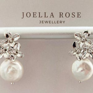 May include: A pair of silver-toned floral earrings with a large, round, white pearl drop. Each earring features three small flowers with clear crystal accents and a pearl that is approximately 1 inch in diameter.