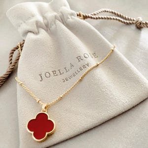 May include: A gold necklace with a red clover pendant is displayed on a light grey jewellery bag. The bag is printed with the text "JOELLA ROSE JEWELLERY". The necklace chain is fine, and the pendant is a four-leaf clover shape.