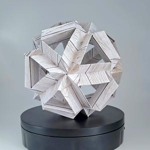 Puede incluir: Una escultura geométrica de papel con un patrón de vetas de madera, exhibida sobre un soporte circular negro. La escultura es una compleja disposición de formas triangulares y rectangulares, creando una forma esférica abierta. El fondo es gris claro liso.