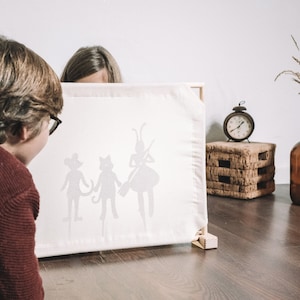 May include: Two children are watching a shadow puppet show. The screen is white and the puppets are black silhouettes of a cat, a mouse, and a fairy. The fairy is holding a wand.