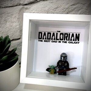 Może przedstawiać: Białe pudełko cieni z czarno-białym nadrukiem, na kt&oacute;rym widnieje napis "The Dadalorian The Best Dad in the Galaxy". Wewnątrz pudełka znajdują się dwie figurki Lego, jedna przedstawiająca Mandalorianina, a druga Grogu.