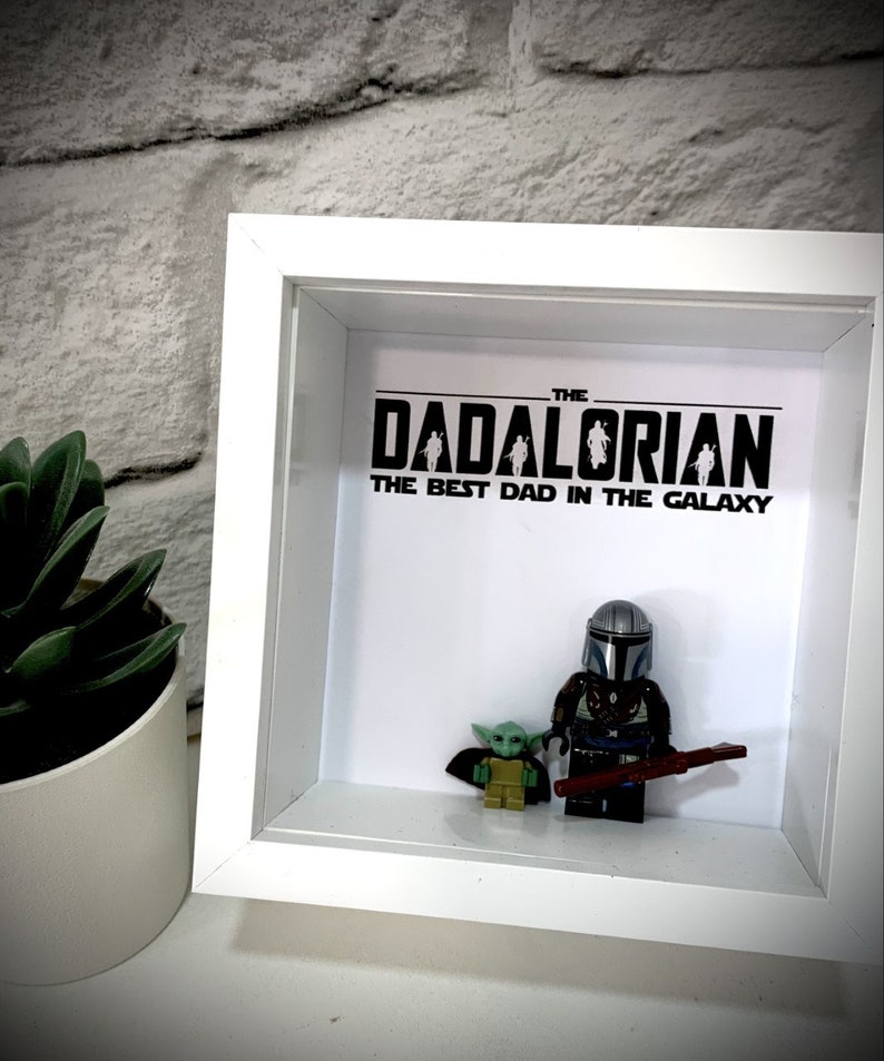 Może przedstawiać: Białe pudełko cieni z czarno-białym nadrukiem, na kt&oacute;rym widnieje napis "The Dadalorian The Best Dad in the Galaxy". Wewnątrz pudełka znajdują się dwie figurki Lego, jedna przedstawiająca Mandalorianina, a druga Grogu.