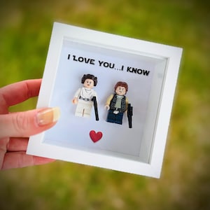 May include: A white framed shadow box featuring Lego figures of Princess Leia and Han Solo, with the text "I LOVE YOU...I KNOW". A red heart is placed below the figures. The figures are holding black accessories.