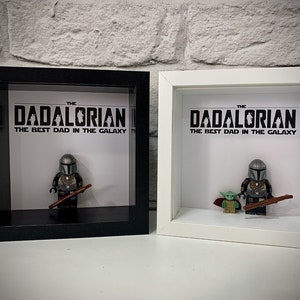 Może przedstawiać: Dwie ramki z cieniowaniem, jedna czarna, a druga biała, każda z nich zawiera minifigurkę Lego Mandalorianina i minifigurkę Grogu. Tekst na białym tle brzmi "THE DADALORIAN THE BEST DAD IN THE GALAXY".