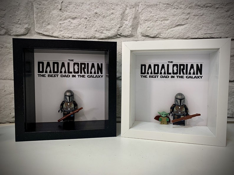 Może przedstawiać: Dwie ramki z cieniowaniem, jedna czarna, a druga biała, każda z nich zawiera minifigurkę Lego Mandalorianina i minifigurkę Grogu. Tekst na białym tle brzmi "THE DADALORIAN THE BEST DAD IN THE GALAXY".