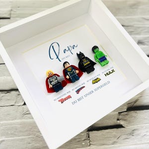 Op de afbeelding: Een ingelijst kunstwerk met Lego-figuren van Thor, Superman, Batman en Hulk. De tekst luidt "Papa, je bent onze superheld". De figuren staan op een witte achtergrond in een witte lijst, een persoonlijk cadeau.