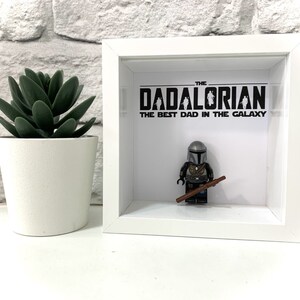 Może przedstawiać: Białe pudełko z cieniowaniem z czarno-białym obrazkiem postaci z Gwiezdnych Wojen, Mandalorianina, z tekstem "The Dadalorian, The Best Dad in the Galaxy".