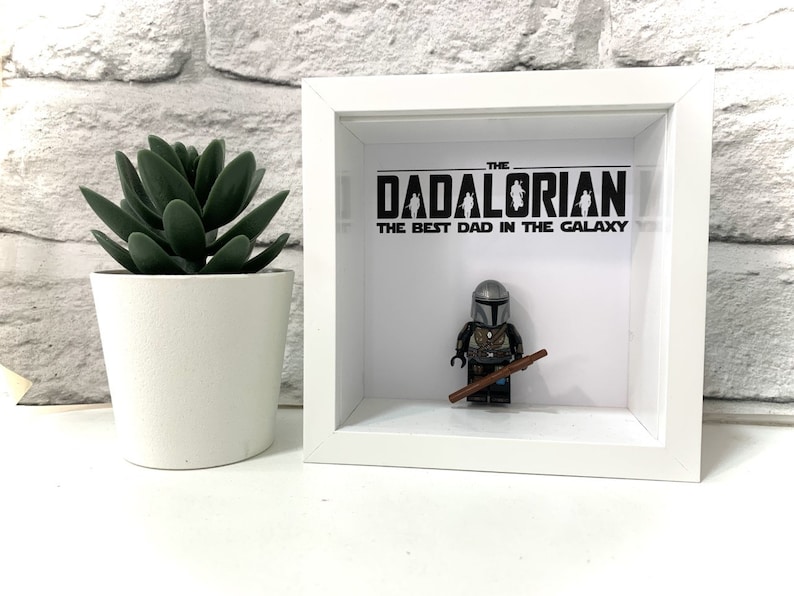 Może przedstawiać: Białe pudełko z cieniowaniem z czarno-białym obrazkiem postaci z Gwiezdnych Wojen, Mandalorianina, z tekstem "The Dadalorian, The Best Dad in the Galaxy".