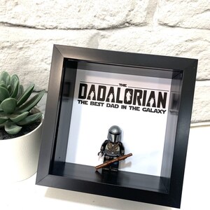 Może przedstawiać: Czarna, oprawiona ramka z cieniowaniem, z białym tłem. Na tle nadrukowany jest tekst "The Dadalorian The Best Dad in the Galaxy". W środku ramki znajduje się figurka Lego Mandaloriana.