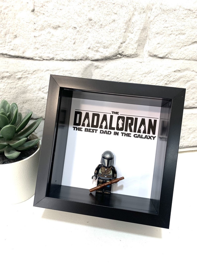 Może przedstawiać: Czarna, oprawiona ramka z cieniowaniem, z białym tłem. Na tle nadrukowany jest tekst "The Dadalorian The Best Dad in the Galaxy". W środku ramki znajduje się figurka Lego Mandaloriana.
