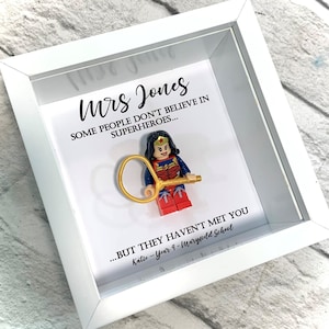 Puede incluir: Un marco blanco con un fondo blanco y una figura de Lego de Wonder Woman sosteniendo un lazo dorado. El texto en el marco dice "Mrs Jones Algunas personas no creen en superhéroes... ...pero no te han conocido Katie - Año 4 - Escuela Maryfield."