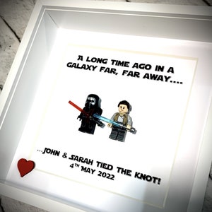 Puede incluir: Un marco blanco con un paspartú blanco y un fondo blanco. El texto "Hace mucho tiempo en una galaxia muy, muy lejana... ...John & Sarah se casaron! 4 de mayo de 2022" está impreso en el fondo. Dos minifiguras de Lego están colocadas en el paspartú. La minifigura de la izquierda es Darth Vader y la minifigura de la derecha es Rey. Un corazón rojo está en la esquina inferior izquierda del marco.
