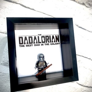 Może przedstawiać: Czarna, oprawiona ramka z cieniowaniem, z białym tłem, na kt&oacute;rym widnieje napis "The Dadalorian, The Best Dad in the Galaxy". W ramce stoi figurka Lego Mandaloriana trzymająca brązową broń.