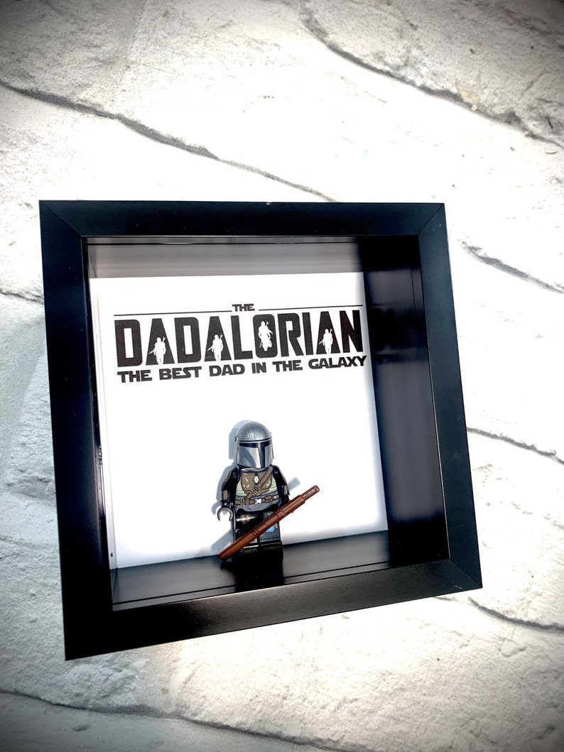 Może przedstawiać: Czarna, oprawiona ramka z cieniowaniem, z białym tłem, na kt&oacute;rym widnieje napis "The Dadalorian, The Best Dad in the Galaxy". W ramce stoi figurka Lego Mandaloriana trzymająca brązową broń.