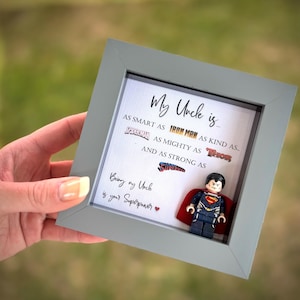 Puede incluir: Una obra de arte enmarcada con el texto "My Uncle is..." y referencias de superhéroes. Una figura de Lego Superman está en el marco. El marco es gris y está sostenido por una mano sobre un fondo verde borroso.
