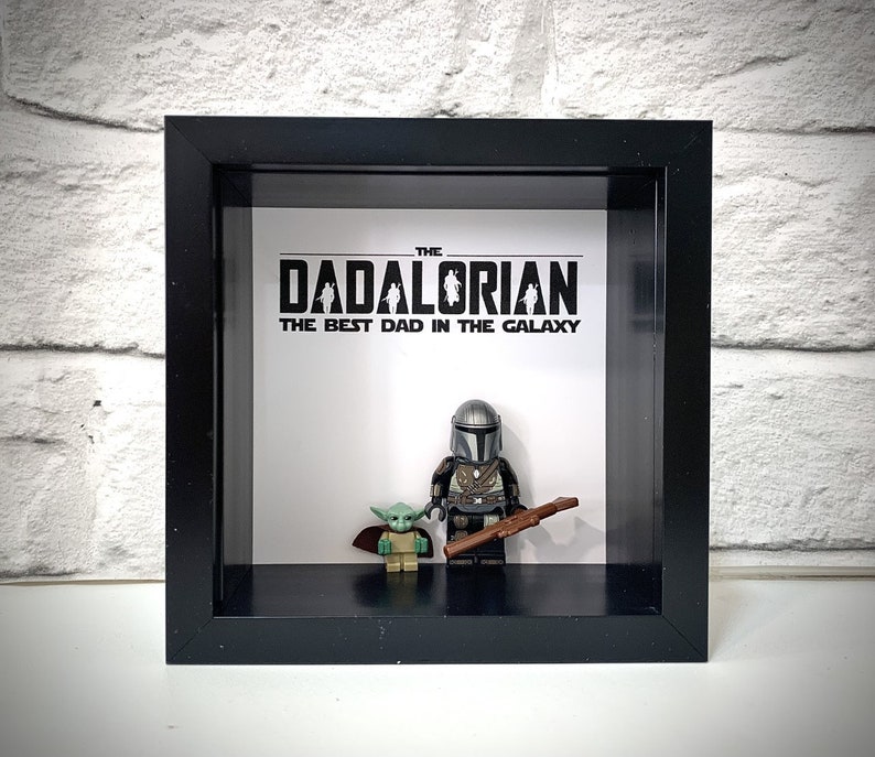 Może przedstawiać: Czarna ramka z cieniowaniem z białym tłem, na kt&oacute;rym widnieje napis "The Dadalorian, The Best Dad in the Galaxy". Wewnątrz ramki znajdują się dwie figurki Lego, jedna przedstawiająca Mandalorianina, a druga Grogu (Baby Yoda).