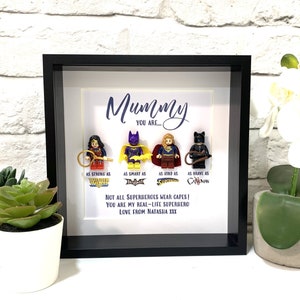 Op de afbeelding: Een ingelijst afdruk met een zwarte lijst en een witte achtergrond. De afdruk bevat een bericht dat luidt "Mummy You Are..." en vier Lego-minifiguren van vrouwelijke superhelden. De tekst onder de minifiguren luidt "Not all superheroes wear capes! You are my real-life superhero Love from Natasha XXX"