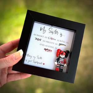 以下が含まれることがあります： 白い背景に黒い文字で「My Sister is... AS SMART AS... AS MIGHTY AS... AND AS COOL AS...」と書かれた額装されたプリント。スーパーヒーローのロゴとハーレイ・クインのレゴミニフィギュアが飾られています。下のテキストには「Being my Sister is your Superpower.」と書いてあります。