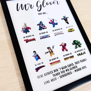 May include: Framed print with a black border featuring superhero illustrations and text. The print includes characters like Thor, Captain America, and Spiderman, with phrases such as "As Mighty As" and "Real Heroes Don't Wear Capes, They Teach!"