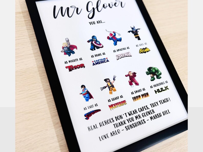 May include: Framed print with a black border featuring superhero illustrations and text. The print includes characters like Thor, Captain America, and Spiderman, with phrases such as "As Mighty As" and "Real Heroes Don't Wear Capes, They Teach!"