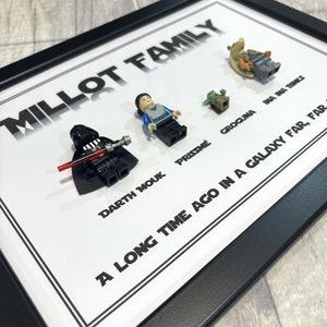 May include: A framed image with a black background and white text. The text reads "Millot Family" and lists names with corresponding Lego minifigures. The minifigures are Darth Vader, a blue-clad figure, a small green figure, and a brown figure.