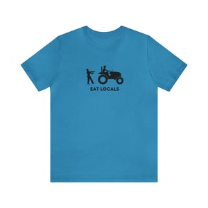 Puede incluir: Una camiseta azul turquesa con una silueta negra de una persona conduciendo un tractor y otra persona corriendo detrás. El texto "EAT LOCALS" está impreso debajo de la imagen.