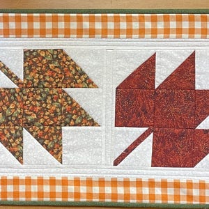 May include: A rectangular fall-themed table runner featuring four stylized leaf designs. The leaves are in shades of yellow, red, and patterned fabrics. The runner has a white center and is bordered by orange and white gingham.