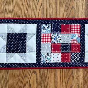 May include: A rectangular quilted table runner with a patriotic theme. The runner features alternating blocks of star and patchwork designs in red, white, and navy blue. The edges are trimmed with a red border.