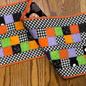 May include: A rectangular quilted table runner featuring a patchwork design of orange, lime green, purple, and black fabric squares with various patterns. The runner is bordered by a black and white gingham check fabric and a bright orange trim.