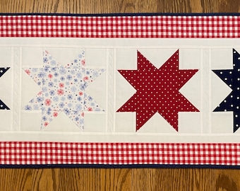 May include: A red, white, and blue patriotic table runner with a gingham border and four star-shaped fabric appliques. The stars are in different patterns, including polka dots, floral, and solid colors.