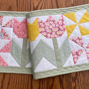 May include: A handmade quilt featuring floral and geometric patterns. The quilt showcases pinwheel and tulip designs in pink, yellow, and white fabrics, with a green border. The quilt is likely used as a table runner or decorative piece.