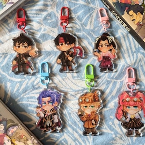 Könnte beinhalten: Sammlung von Acryl-Schlüsselanhängern mit Anime-Charakteren mit verschiedenen Outfits und Accessoires. Jeder Schlüsselanhänger hat einen unterschiedlich farbigen Verschluss. Die Schlüsselanhänger sind auf einer blau gemusterten Oberfläche angeordnet, mit einem Buch im Hintergrund.