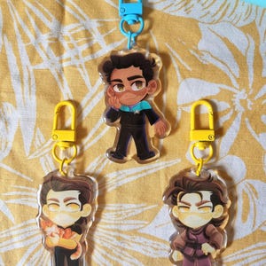 May include: Three acrylic keychains featuring cartoon characters. One keychain has a blue clasp, while the other two have yellow clasps. The characters are depicted in various outfits and poses, with detailed facial features and expressions.