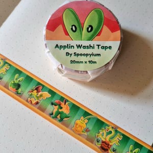 Peut inclure: Un rouleau de ruban adhésif Washi Applin par Spoopyium, 20 mm x 10 m. Le ruban présente un motif répété de personnages de dessins animés verts, jaunes et oranges sur un fond à rayures vertes et dorées. Le rouleau a un design rouge et orange avec des yeux verts.
