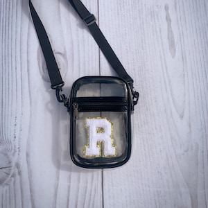 May include: Clear crossbody bag with a black strap and zipper. The bag features a white letter "R" patch with gold glitter accents. The bag is designed to be worn over the shoulder.