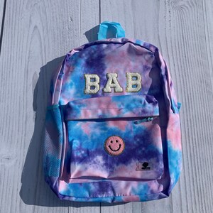 Peut inclure: Un sac à dos tie-dye dans les tons de bleu, violet et rose. Le sac à dos porte les lettres "BAB" en or, une poche avant zippée et un écusson smiley. Il a une poignée bleue et une poche latérale en filet. Mesures en cm.
