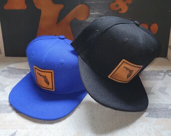 May include: Two baseball caps, one royal blue and one black, are displayed. Each cap features a square leather patch with the outline of Florida. The caps have a flat brim and a structured crown. The background includes a wooden sign.