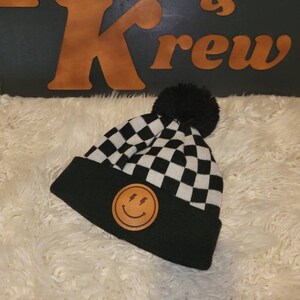 May include: Black and white checkered beanie with a black pom pom on top. The beanie has a brown leather patch with a smiley face and lightning bolts on it.